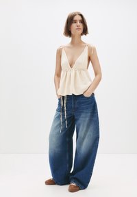 Ung kvinna står med händerna i fickorna, iförd en krämfärgad stickad ärmlös topp och vida, bleka blå jeans med bruna skor.