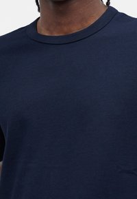 Άνω μέρος σώματος ατόμου που φοράει απλό σκούρο μπλε μπλούζα με στρογγυλή λαιμόκοψη σε λευκό φόντο.