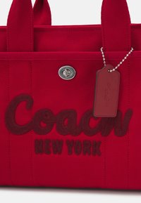 Sac fourre-tout en tissu rouge avec "Coach New York" brodé, fermeture à verrou argentée et une étiquette rouge assortie sur une chaîne attachée à la poignée.