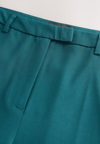 Pantalones de color verde azulado con una textura suave, diseño de frente plano, dos pinzas y cinturilla con trabillas para cinturón. Se observan detalles de costura.