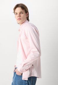 Cămașă roz deschis cu dungi, dintr-un material moale, având guler cu nasturi, mâneci lungi și o croială lejeră cu slituri laterale și un accent cu logo.