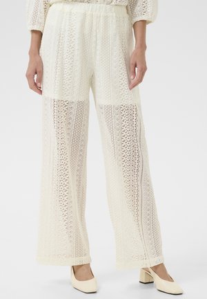 Fehér csipkés, bő szárú nadrág finom mintákkal, állítható derékkal és légies sziluettel, bézs blokk-sarkú cipővel párosítva.