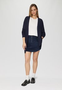 Navy ribbed cardigan gedragen over een wit T-shirt, gecombineerd met een navy corduroy rok met een voorrits, en zwarte gepunte loafers.
