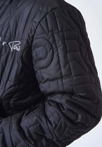 Gros plan sur une veste matelassée noire avec des coutures géométriques et un texte brodé blanc « "Paris" » sur la poitrine.