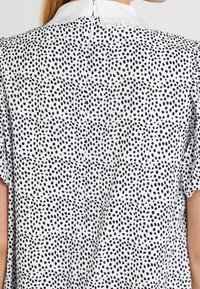 Black and white polka dot blouse with a stand-up collar, short sleeves, and a button closure at the back. Smooth fabric texture.