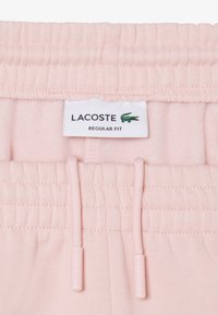 Ceinture élastique rose clair avec cordons sur un vêtement, avec une étiquette blanche Lacoste arborant un logo crocodile vert et le texte "Regular Fit".