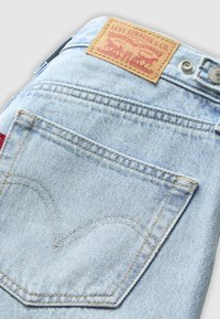 Jeans in denim blu chiaro con un design distintivo della tasca posteriore caratterizzato da un arco cucito. Cuciture dorate e un'etichetta in pelle color beige sono visibili.
