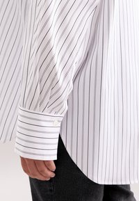 Camicia bianca a maniche lunghe a righe con sottili righe nere, dotata di polsini con bottone e un orlo curvo, realizzata in tessuto morbido.