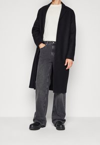 Un long manteau noir par-dessus une chemise claire, associé à un jean large gris. Des bottes noires confortables complètent la tenue, avec une texture lisse.
