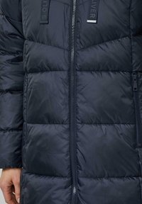 Veste matelassée bleu marine avec un design matelassé, des fermetures éclair apparentes et des accents de logo. Fabriquée en matériau lisse et résistant à l'eau.