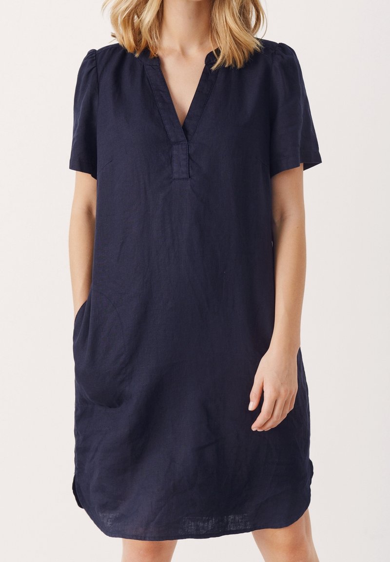 Femme portant une robe bleu marine ample, à manches courtes, longueur genou, avec un col en V et des poches latérales, debout devant un fond uni.