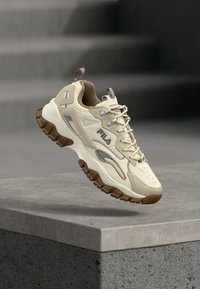 Μπεζ αθλητικό παπούτσι με υφασμάτινο επάνω μέρος, κρεμ δερμάτινες λεπτομέρειες, καφέ λαστιχένια σόλα και κορδόνια. Διαθέτει λογότυπο στο πλάι και στη γλώσσα της φτέρνας.