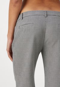 Gros plan d'une personne portant un pantalon à carreaux gris et noir montrant les poches arrière et une partie du bas du dos et du bras nus.