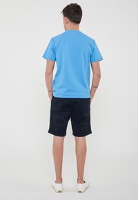 T-shirt in cotone blu con maniche corte, abbinato a pantaloni corti di un blu navy scuro. Il modello indossa scarpe slip-on bianche. Design minimalista, senza motivi visibili.