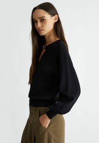 Maglione nero a maglia con scollatura a V e un distintivo dettaglio in metallo. Maniche lunghe a palloncino con polsini a coste, abbinato a pantaloni verde oliva.
