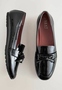 Mocassini in vernice nera con punta rotonda, dettaglio decorativo a fiocco e accenti testurizzati. L'interno presenta una fodera bordeaux. Suola resistente.