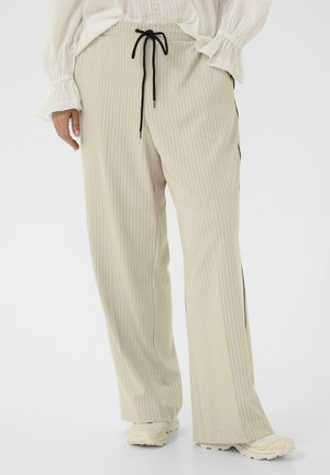 Persoon draagt beige pantalon met wijde pijpen en beige krijtstreep, zwarte trekkoord, witte sneakers en een wit blouse met lange mouwen en kanten manchetten.