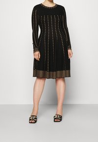 Robe noire à manches longues avec motifs en diamants dorés, dotée d'un col rond et d'une jupe évasée. Associée à des sandales plates noires avec des accents dorés.