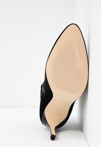 Chaussure à talon haut en daim noir avec un bout pointu et un talon stiletto fin. La semelle est beige avec une texture nervurée et le logo de la marque embossé.