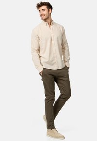 Chemise beige clair à boutons avec manches longues, associée à un pantalon slim vert olive et des chaussures slip-on de couleur claire.