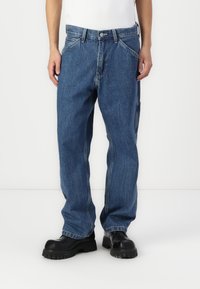 Homme portant un jean bleu large et des bottes noires épaisses, debout devant un fond blanc uni.