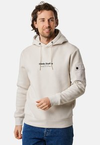 Felpa crema con cappuccio e lacci, con un logo sul davanti. Presenta un tessuto a trama, polsini a coste e una tasca sulla manica.