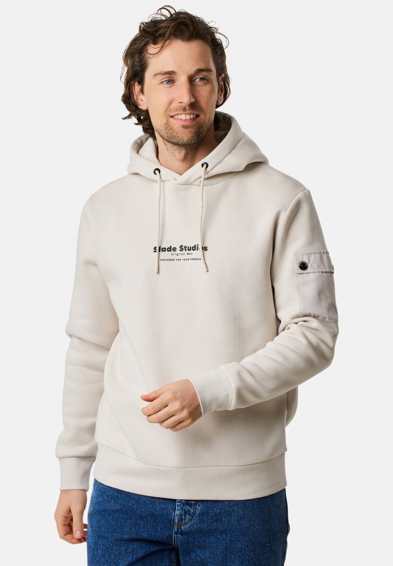 Felpa crema con cappuccio e lacci, con un logo sul davanti. Presenta un tessuto a trama, polsini a coste e una tasca sulla manica.
