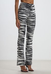 Pantaloni a zebrato bianco e nero a campana con vita alta, texture liscia e chiusura frontale a bottoni, abbinati a sandali neri con tacco.