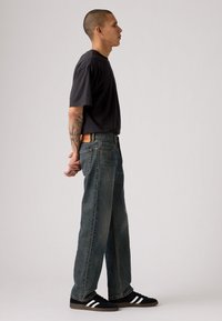 Camiseta negra de cuello redondo; jeans de pierna ancha azules, ligeramente desgastados; zapatillas negras con rayas blancas. Accesorios mínimos, vista de perfil lateral.