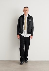 Veste matelassée noire avec un écusson de logo, chemise blanche, cravate beige, pantalon noir et chaussures noires. Le mannequin se tient devant un mur uni.