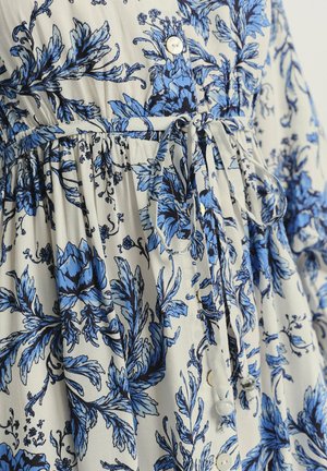 Vestido floral en beige claro con patrones botánicos azules. Cuenta con una cintura ajustada, lazo de tela y botones redondos blancos. Material suave y ligero.