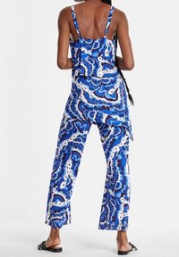 Blå mönstrad jumpsuit med spaghetti-stroppar, vida ben och löst sittande passform. Har en virvlande design i olika nyanser av blått och vitt.