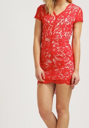 Nainen pukeutuneena lyhyeen, punaiseen, kukalliseen pitsimekkoon, jossa on lyhyet hihat ja V-kaula-aukko, seisoo jalka hieman koukistettuna.