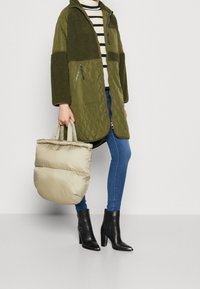 Person som bär en grön vadderad och fleecejacka, blå slim fit jeans, svarta klackstövletter och håller en beige vadderad handväska.