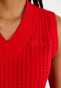 Rood gebreide mouwloze top met een V-hals, een gestructureerd open breipatroon en een geborduurd logo-detail op de linkerzijde van de voorkant.