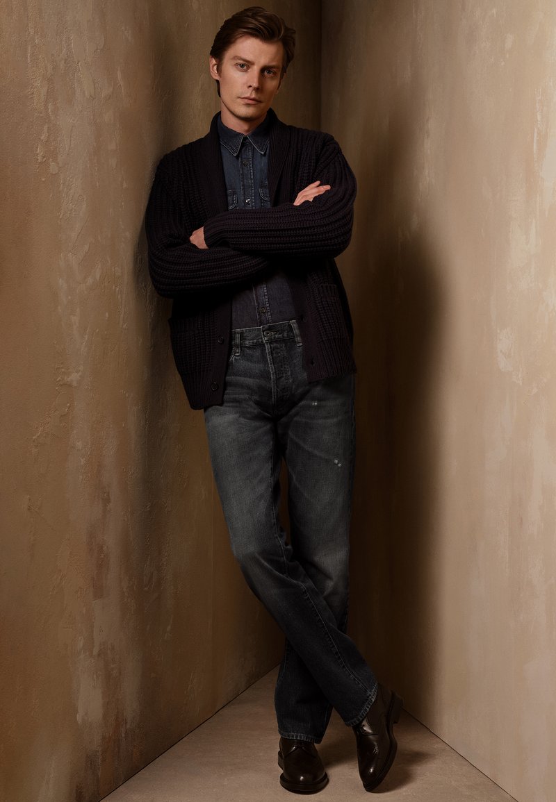 Tamno pletivo kardigan preko traper košulje, uparen sa sivim farmerkama i crnim cipelama. Subjekt stoji naslonjen na teksturirani, neutralni zid.