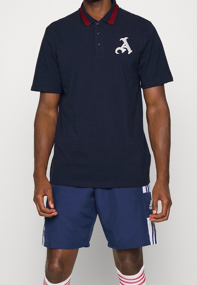 Homme portant un polo bleu marine avec des rayures rouges sur le col et une lettre A blanche, un short Adidas bleu avec des rayures blanches, et des chaussettes blanches à rayures rouges.