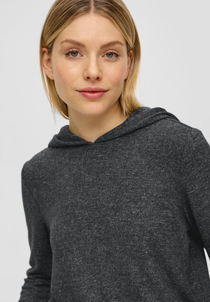 Junge Frau mit hellblondem Haar, die einen dunkelgrauen Kapuzenpullover trägt, Gesicht nach vorn gewandt und mit einem leichten Lächeln vor einem einfarbigen, hellen Hintergrund.