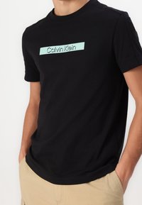 Schwarzes Baumwoll-T-Shirt mit einem hellblauen "Calvin Klein"-Logo in rechteckigem Design. Kurze Ärmel und Rundhalsausschnitt.