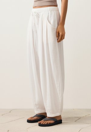 Femme portant un pantalon ample en lin blanc avec poches et des tongs compensées noires, debout sur un sol carrelé.