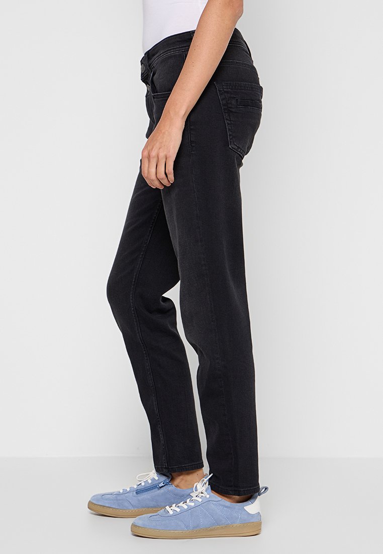 Osoba ubrana w czarne dżinsy o prostym kroju i jasnoniebieskie zamszowe trampek stoi na tle białym.