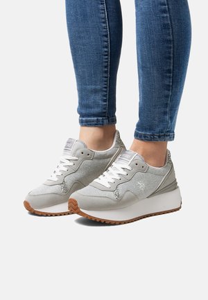 Pieds portant des baskets argentées pailletées avec lacets blancs et semelles en gomme, assortis à un jean skinny bleu sur fond blanc.