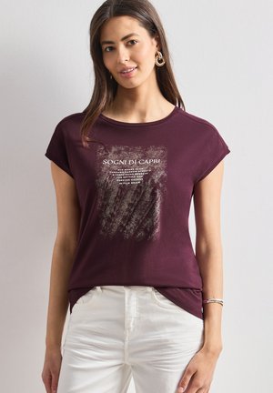 Kvinde med langt mørkt hår iført gyldne hoops-øreringe, en bordeaux T-shirt med hvid tekst og hvide bukser, stående mod en ensfarvet baggrund.