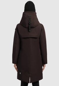 Abrigo aislante marrón con un cuello alto, correa ajustable y panel de ventilación en la espalda. Cuenta con un parche de logotipo cerca del dobladillo y un corte recto.
