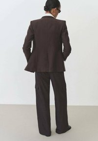 Bruine op maat gemaakte blazer met een contrasterende witte kraag, gecombineerd met losse bruine cargobroeken met zijzakken. Zwarte schoenen maken de outfit compleet.