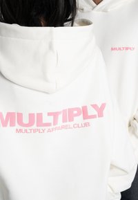 Sudadera blanca hecha de una tela suave con el texto rosa "MULTIPLY" en la parte posterior. Presenta una gran capucha y un corte relajado, adecuada para un uso casual.