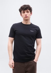 Νεαρός άνδρας με σγουρά σκούρα μαλλιά, φοράει μαύρο μπλουζάκι Pepe Jeans και καφέ παντελόνι, στέκεται με το ένα χέρι στην τσέπη μπροστά σε απλό φόντο.