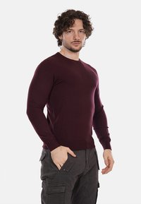 Suéter de punto burdeos con mangas largas y cuello redondo, diseño ajustado, textura suave. Combinado con pantalones cargo oscuros.
