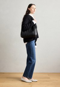 Svart plisserad handväska med en slät yta, kombinerad med en svart stickad tröja, blå jeans och vita platta skor på ett trägolv.