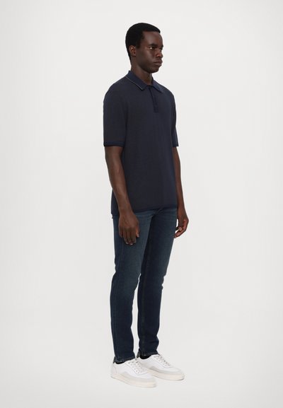 Mørkeblå polo med kort erme, ribbet tekstur og knappestolpe. Brukt med mørke jeans og hvite joggesko.